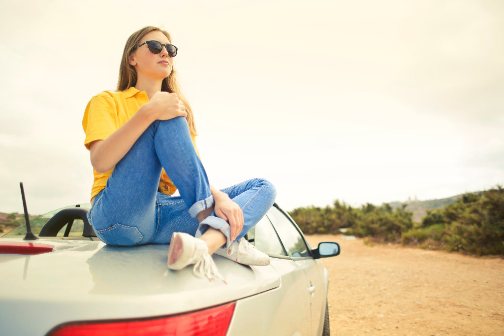 Młoda dziewczyna w okularach przeciwsłonecznych, żółtej koszuli i jeansach siedząca na samochodzie